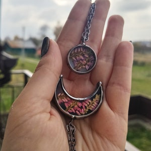 May include: A silver chain necklace with two pendants. The larger pendant is a crescent moon shape with dried pink flowers inside. The smaller pendant is a circle with dried pink flowers inside.