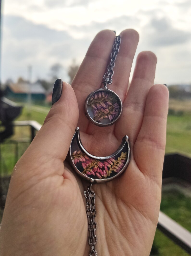 May include: A silver chain necklace with two pendants. The larger pendant is a crescent moon shape with dried pink flowers inside. The smaller pendant is a circle with dried pink flowers inside.