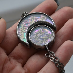 May include: A silver necklace with a double crescent moon pendant. The pendant is filled with pressed flowers in shades of pink and purple.
