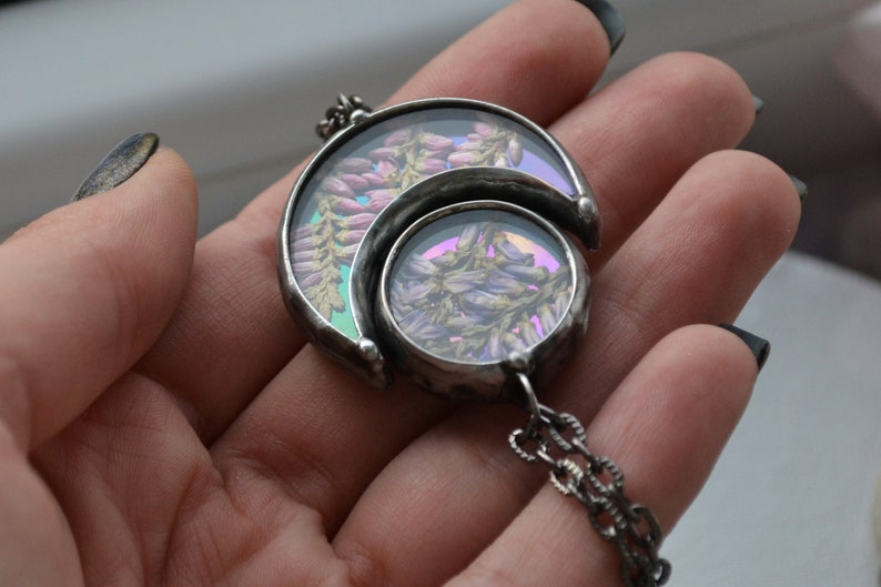 May include: A silver necklace with a double crescent moon pendant. The pendant is filled with pressed flowers in shades of pink and purple.