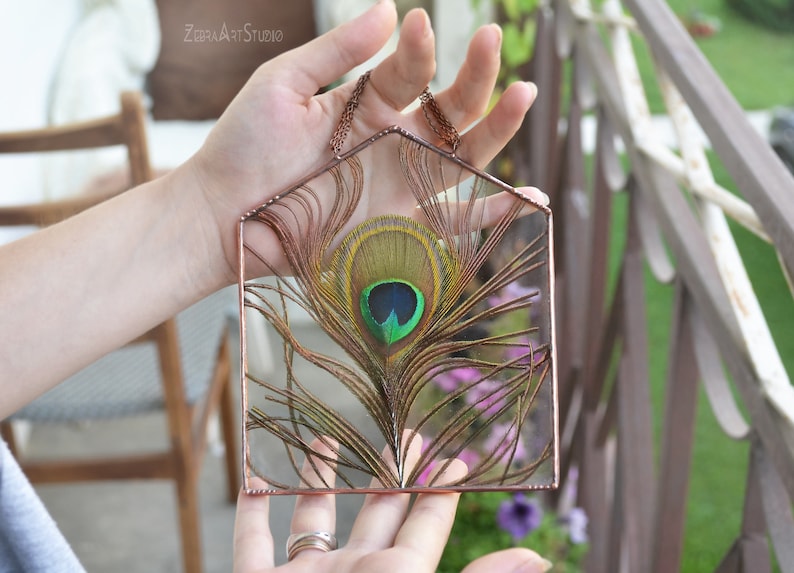 May include: A copper wire frame with a peacock feather inside. The feather is brown, green, and blue with a large eye spot. The frame is hanging from a copper chain.