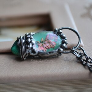 May include: A silver pendant necklace with a green and pink resin oval charm featuring pressed flowers. The pendant is attached to a silver chain.