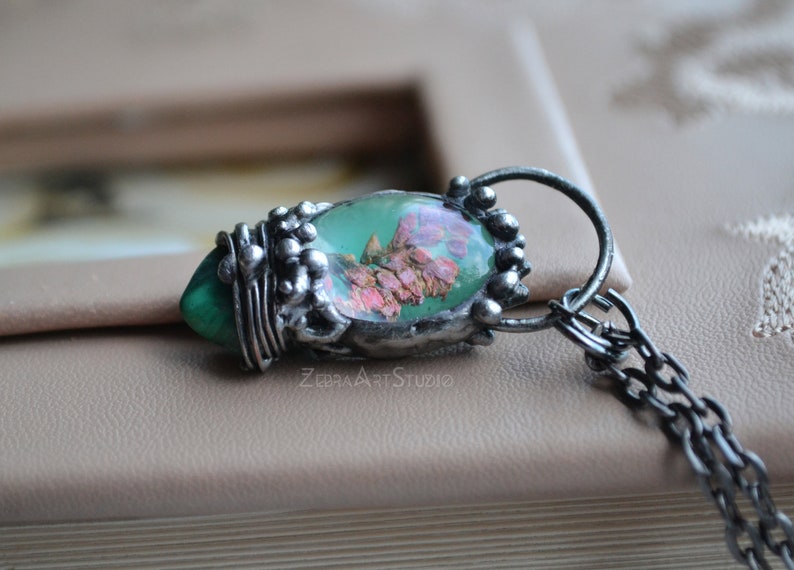 May include: A silver pendant necklace with a green and pink resin oval charm featuring pressed flowers. The pendant is attached to a silver chain.