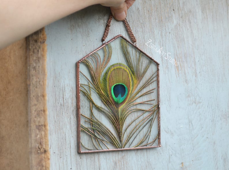 May include: A copper-framed glass window with a peacock feather inside. The feather is green, blue, and gold with a large eye spot.