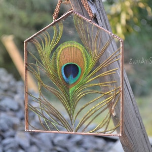 May include: A copper-framed glass windowpane with a peacock feather inside. The feather is green, blue, and gold with a large eye in the center. The windowpane is hanging from a copper chain.