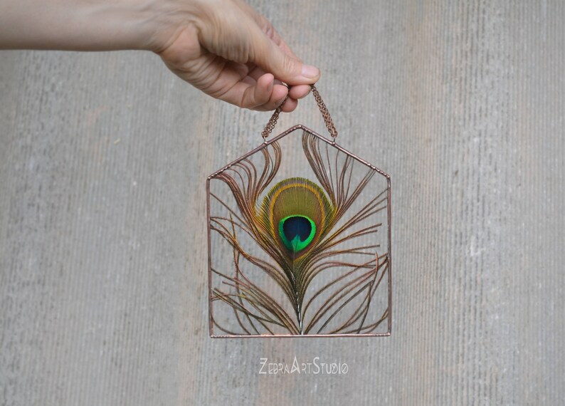 May include: A copper wire frame in the shape of a house with a peacock feather inside. The feather is green, blue, and gold. The frame is hanging from a copper chain. The text "ZEBRA ART STUDIO" is at the bottom of the image.
