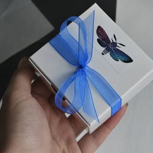 May include: A white gift box with a blue ribbon and a butterfly design. The butterfly is iridescent and has black, blue, and purple wings.