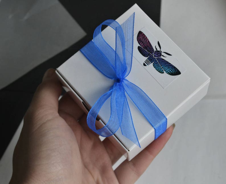 May include: A white gift box with a blue ribbon and a butterfly design. The butterfly is iridescent and has black, blue, and purple wings.
