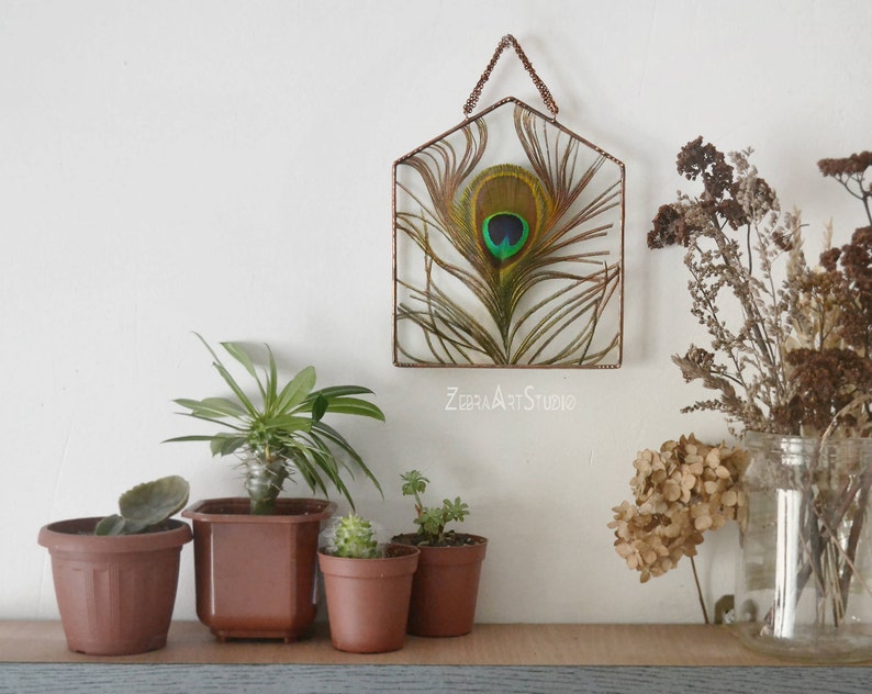 May include: A copper wire house-shaped frame with a preserved peacock feather inside. The frame is hanging on a white wall with plants and dried flowers in front of it. The text "ZEBRA ART STUDIO" is visible below the frame.