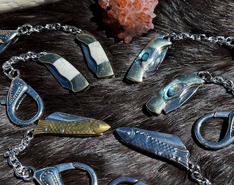 May include: A silver chain with multiple small pocket knives attached. The knives have different designs, including a fish, a shell, and a white and black pattern.