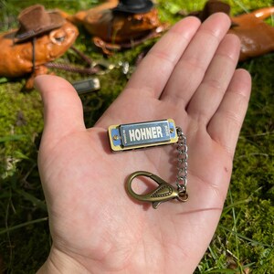 May include: A small silver harmonica keychain with a gold trim and a silver chain. The harmonica has the word "HOHNER" printed on it in black letters. The keychain is held in a hand against a green mossy background.