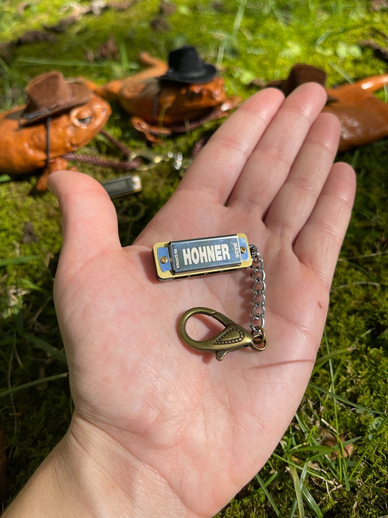 May include: A small silver harmonica keychain with a gold trim and a silver chain. The harmonica has the word "HOHNER" printed on it in black letters. The keychain is held in a hand against a green mossy background.