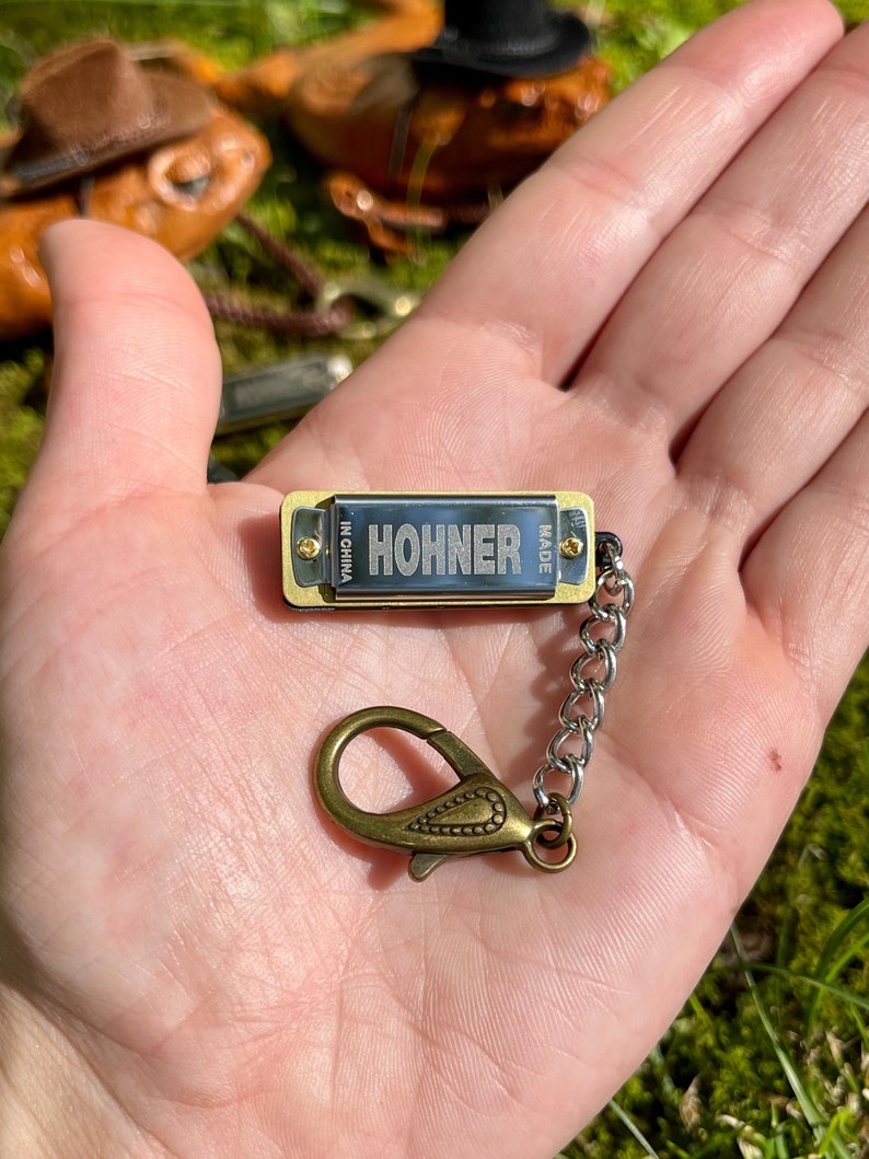May include: A small gold harmonica keychain with the word "HOHNER" printed on it. The harmonica is attached to a silver chain with a gold clasp.