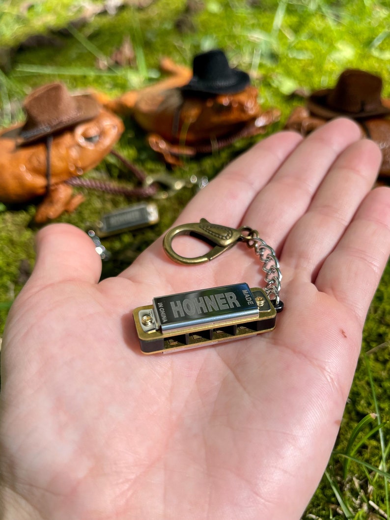 May include: A small gold harmonica keychain with the word "HOHNER" and "MADE IN CHINA" printed on it. The harmonica is attached to a gold chain with a gold clasp.