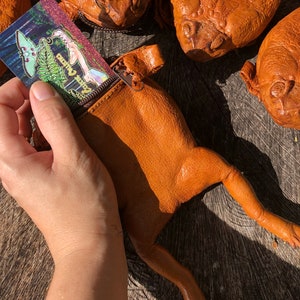 May include: A brown leather frog-shaped purse with a zipper closure. The purse is open and a business card with a colorful design is partially inserted into the zippered pocket. The purse is lying on a wooden surface.