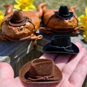 May include: Three miniature cowboy hats, one brown, one black, and one black with a brown band. The hats are on a wooden surface and a hand is holding the brown hat.