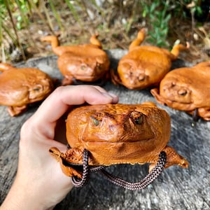 May include: Four brown leather frog-shaped purses. One frog purse is being held in a hand. The frog purses have a textured leather surface and have a brown cord attached to them.