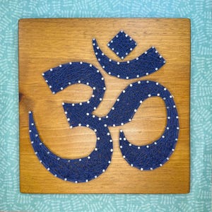 May include: A wooden wall hanging featuring the Om symbol crafted with navy blue string and white nails. The symbol is centered on a rectangular, light brown wooden board. The background is a light blue patterned fabric.
