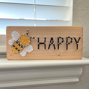 May include: Wooden sign with a string art bee and the word "HAPPY". The bee is yellow, black, and white, while the letters are black. The sign is made of light-colored wood and is displayed on a white surface.