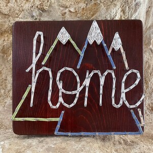 May include: A wooden sign with the word "home" spelled out in white string. The sign features mountain designs in green, blue, and white string. The background is a dark brown wood.