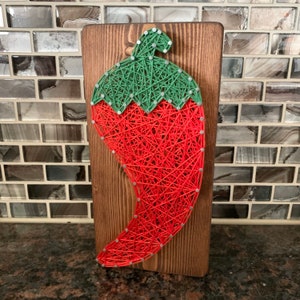 May include: A handmade string art piece featuring a red chili pepper with a green stem, mounted on a stained wooden board. The chili pepper is crafted with red string, and the stem is made with green string. The nails are silver.