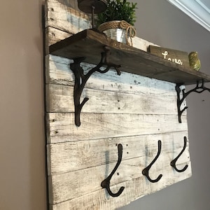 May include: Whitewashed wooden shelf with three black metal hooks and two decorative metal brackets. The shelf has a rustic look and is decorated with a small potted plant and a birdcage.