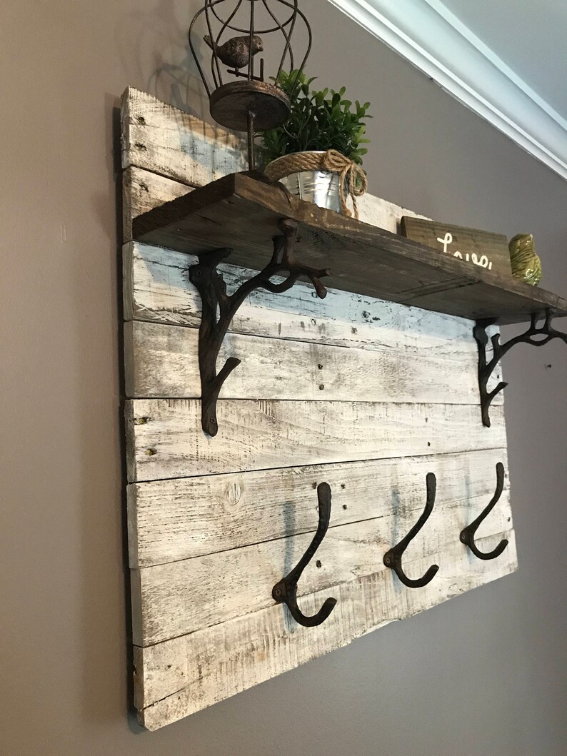 May include: Whitewashed wooden shelf with three black metal hooks and two decorative metal brackets. The shelf has a rustic look and is decorated with a small potted plant and a birdcage.