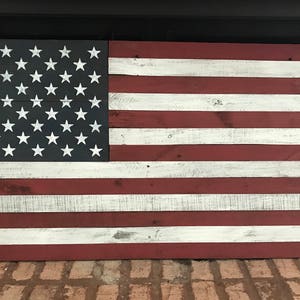 May include: A rustic wooden American flag with red, white, and blue paint. The flag is made of reclaimed wood and has a distressed look. The stars are white and the stripes are red and white.