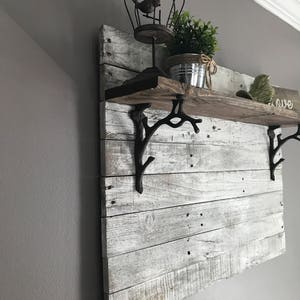 May include: A whitewashed wooden shelf with black metal branch-shaped brackets. The shelf is decorated with a birdcage, a potted plant, and a small decorative bird. The word "love" is written on the wall behind the shelf.