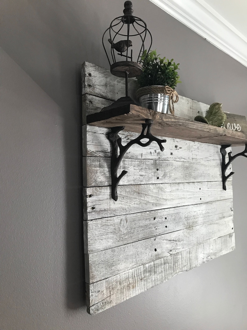 May include: A whitewashed wooden shelf with black metal branch-shaped brackets. The shelf is decorated with a birdcage, a potted plant, and a small decorative bird. The word "love" is written on the wall behind the shelf.