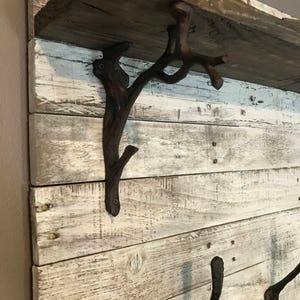 May include: A rustic wooden shelf with whitewashed wood and a distressed finish. The shelf is supported by two metal hooks and a decorative metal branch-shaped bracket. The shelf is perfect for displaying plants, books, or other decorative items.