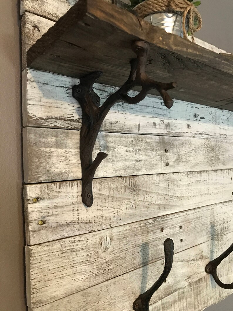 May include: A rustic wooden shelf with whitewashed wood and a distressed finish. The shelf is supported by two metal hooks and a decorative metal branch-shaped bracket. The shelf is perfect for displaying plants, books, or other decorative items.