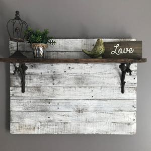 May include: Whitewashed wooden shelf with black metal brackets. The shelf is decorated with a small potted plant, a bird figurine, and a sign that says "Love".