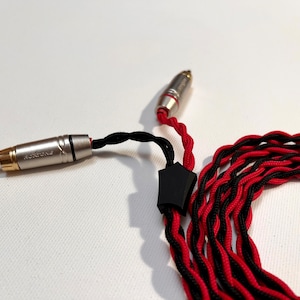 Puede incluir: Un cable de audio trenzado negro y rojo con conectores chapados en oro. El cable tiene un alivio de tensión de plástico negro y está enrollado.