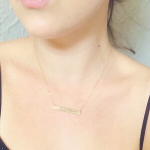 May include: A gold-colored bar necklace with a delicate chain. The rectangular pendant features engraved text. The necklace is worn against a person's skin, with a black top visible.
