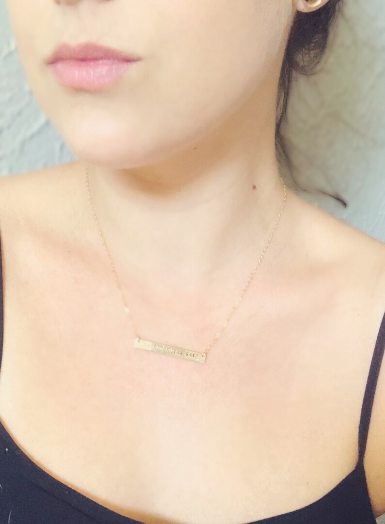 May include: A gold-colored bar necklace with a delicate chain. The rectangular pendant features engraved text. The necklace is worn against a person's skin, with a black top visible.