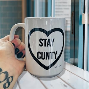 May include: White ceramic mug with a black heart design that says "Stay Cunty" in all caps. The mug has a black handle and the text "Lux + Luca" is printed on the bottom.