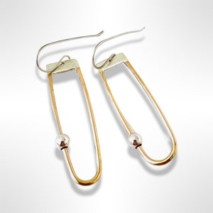 May include: A pair of gold and silver earrings with a simple, geometric design. Each earring features a long, oval-shaped loop with a small silver ball at the center.