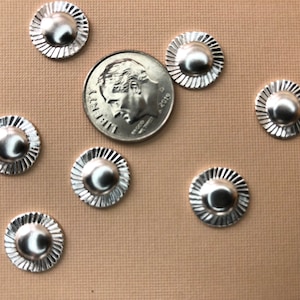 Op de afbeelding: Zeven zilverkleurige metalen versieringen met een verhoogd midden en een decoratieve, gegolfde rand. De versieringen zijn gerangschikt op een lichtbruin oppervlak, met een Amerikaanse dime-munt als maatstaf.
