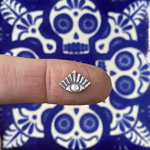 May include: A silver charm in the shape of a sun with a raised center. The charm is on a finger against a background of blue and white tiles with a skull design.