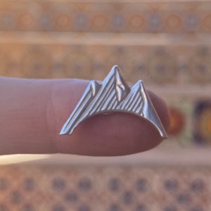 Könnte beinhalten: Eine silberne Bergkette-Anstecknadel mit strukturierter Oberfläche. Die Anstecknadel ist in Form einer Bergkette mit drei Gipfeln gestaltet.