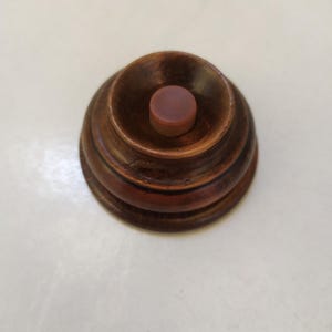 Könnte beinhalten: Kleines, gedrechseltes Holzobjekt mit dunkelbraunem Finish. Das Objekt hat einen abgestuften, abgerundeten Sockel und einen zentralen, kreisförmigen, rotbraunen Verschluss. Das Design deutet auf einen dekorativen oder funktionalen Gegenstand hin, möglicherweise einen Flaschenverschluss.
