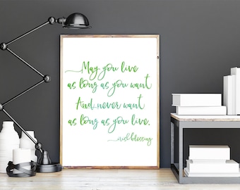 Op de afbeelding: Een ingelijste print met de tekst "May you live as long as you want and never want as long as you live. Irish Blessing" in groen schrift. De print wordt weergegeven op een houten oppervlak met een zwarte bureaulamp en witte decoratie.
