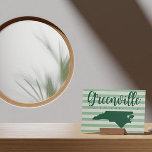 Puede incluir: Tarjeta a rayas verdes y blancas con el texto "Greenville, Carolina del Norte" y un contorno verde del estado de Carolina del Norte con un corazón verde en el centro.