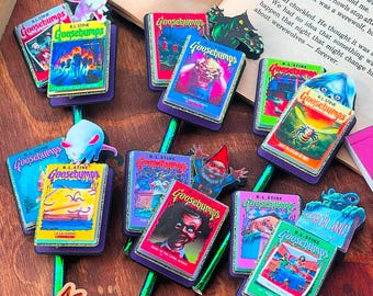 Peut inclure: Une collection de neuf couvertures de livres Goosebumps mettant en scène divers personnages et scènes effrayants. Les couvertures sont attachées à des crayons avec des accents violets et verts. Les crayons sont disposés sur une surface en bois avec les mots "My Most" visibles au premier plan.
