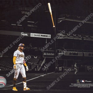 May include: A baseball player in a white and gold striped uniform with the word "PADRE" on the front, stands on a baseball field. A baseball bat is flying through the air above him. The background is a black and white image of a baseball stadium.
