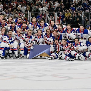 May include: A group of hockey players in white, blue, and red uniforms, with the word "USA" on the front, pose for a photo on an ice rink. Many are wearing gold medals. The crowd in the background is blurred.