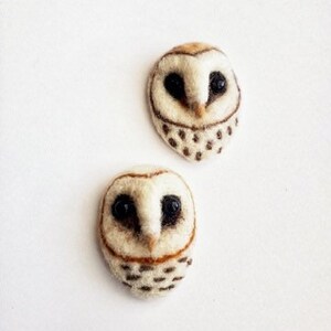 May include: Two handmade owl-shaped brooches. The owls are crafted from off-white felt with brown and black details. The eyes are black, and the lower portion of the owl features small, dark brown spots. The brooches are set against a white background.