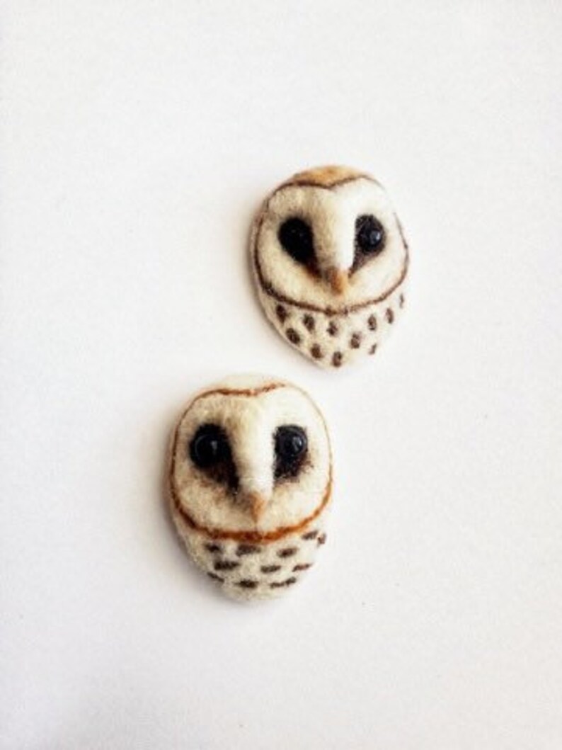 May include: Two handmade owl-shaped brooches. The owls are crafted from off-white felt with brown and black details. The eyes are black, and the lower portion of the owl features small, dark brown spots. The brooches are set against a white background.