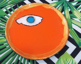 May include: Orange ceramic dish with a painted eye design. The eye is white with a blue pupil and black outline.